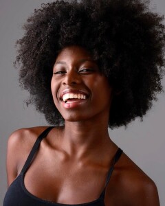 Foto de uma jovem negra sorrindo para a câmera. Ela está de cabelo solto e usa uma blusinha regata preta.
