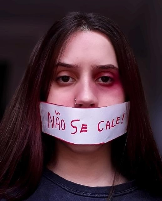 Fotografia de uma jovem com uma faixa branca tampando sua boca. Nela está escrito, em vermelho, "Não se cale!"