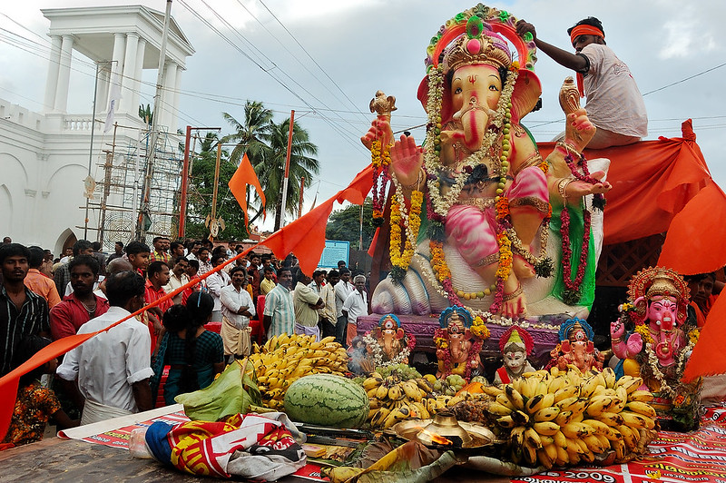 Foto de uma estátua da divindade Ganesha repleta de furtas e outros alimentos. Há pessoas em volta