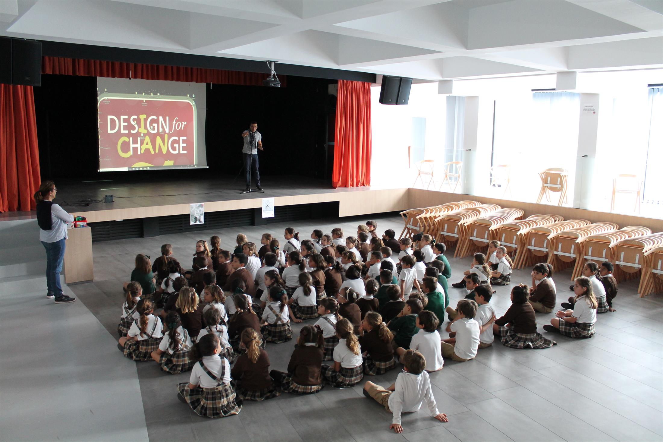 Foto de fundo de um salão, em que há crianças sentadas enfileiradas assistindo a um telão com o logo do Design For Change.